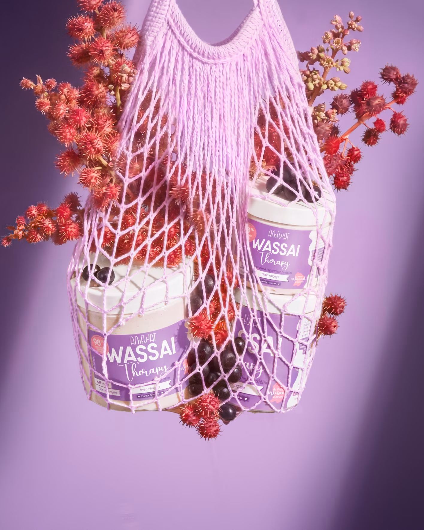 Amewat Wassaï Therapy jars suspended inside a lilac market net with açai berries and red castor flowers against a purple studio background.
