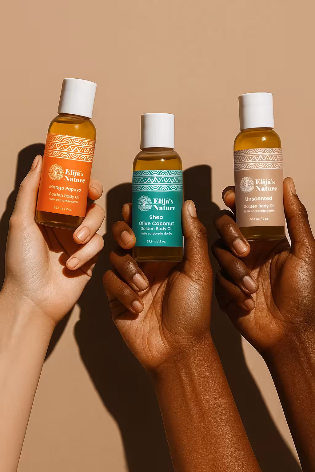 Three hands of different skin tones holding Elija’s Nature mini body oils: Mango Papaya, Olive Coconut, and Unscented against a beige backdrop.