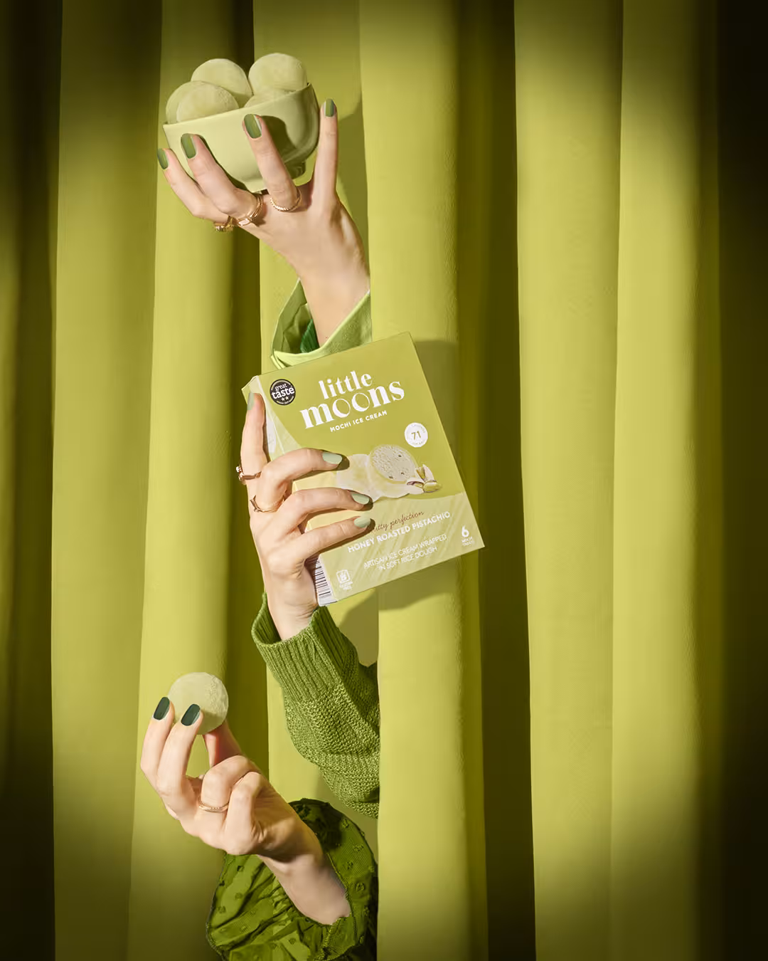 Hands emerging from green curtains holding Little Moons Pistachio mochi and packaging.