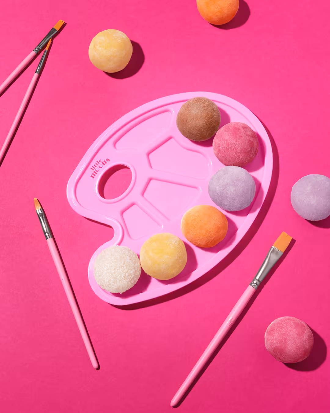 Little Moons mochi arranged in a painter’s palette with paintbrushes on bold pink backdrop.