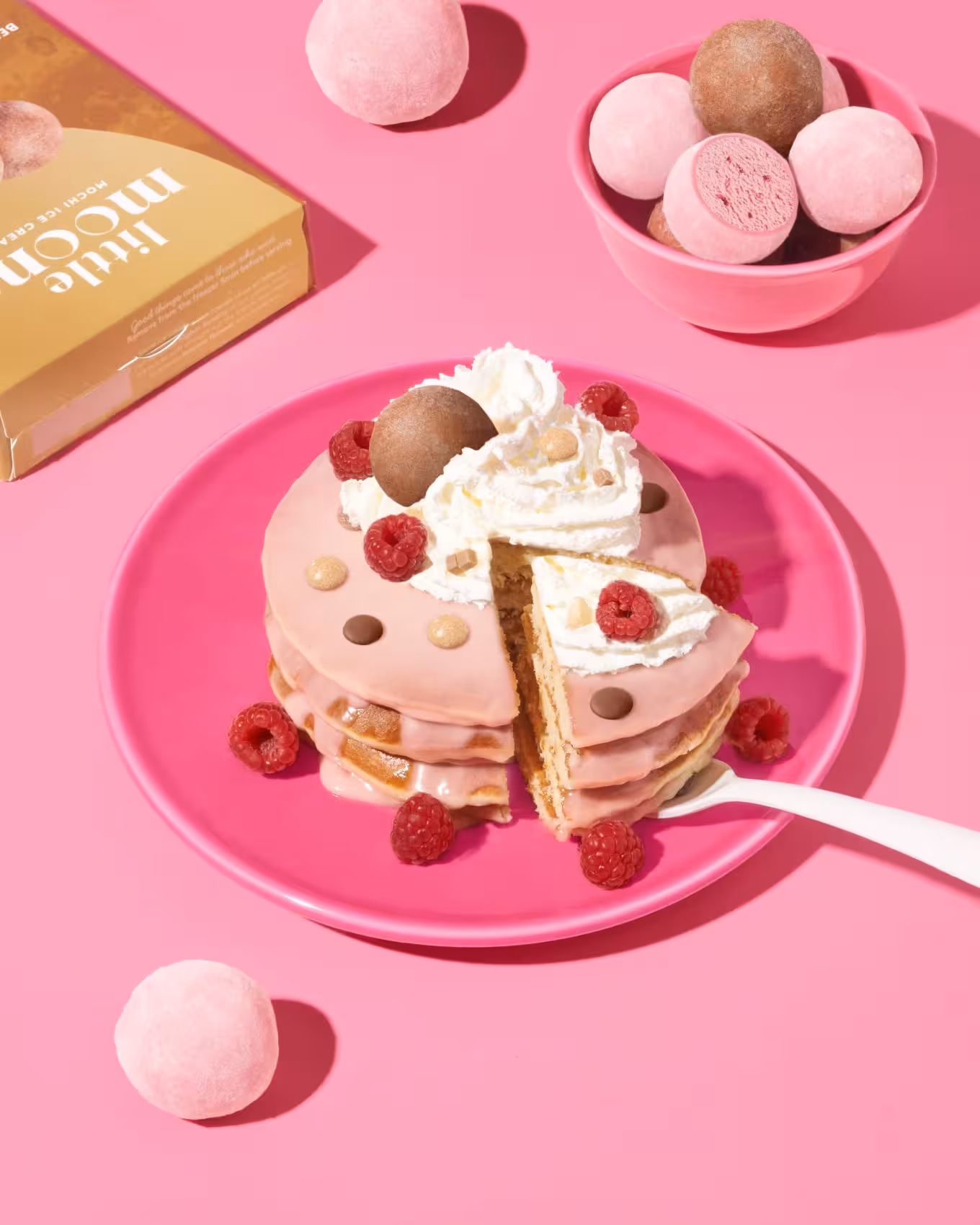 Stack of pancakes topped with Little Moons mochis, whipped cream, and raspberries on pink plate.