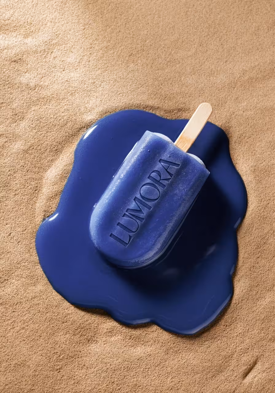Creative campaign image of a Lumora-branded blue popsicle melting on sand.