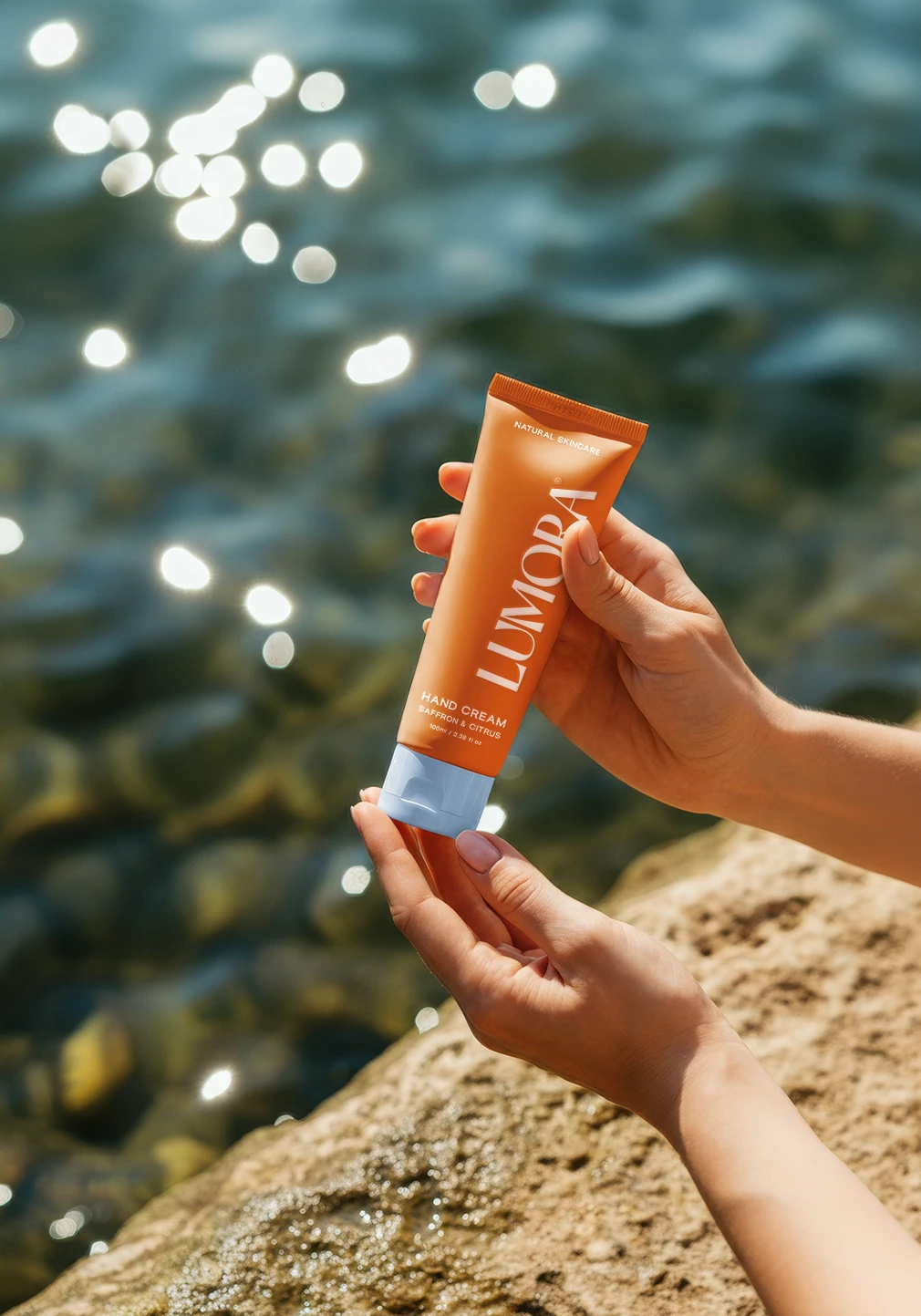 Hands holding Lumora saffron and citrus hand cream tube by the water.