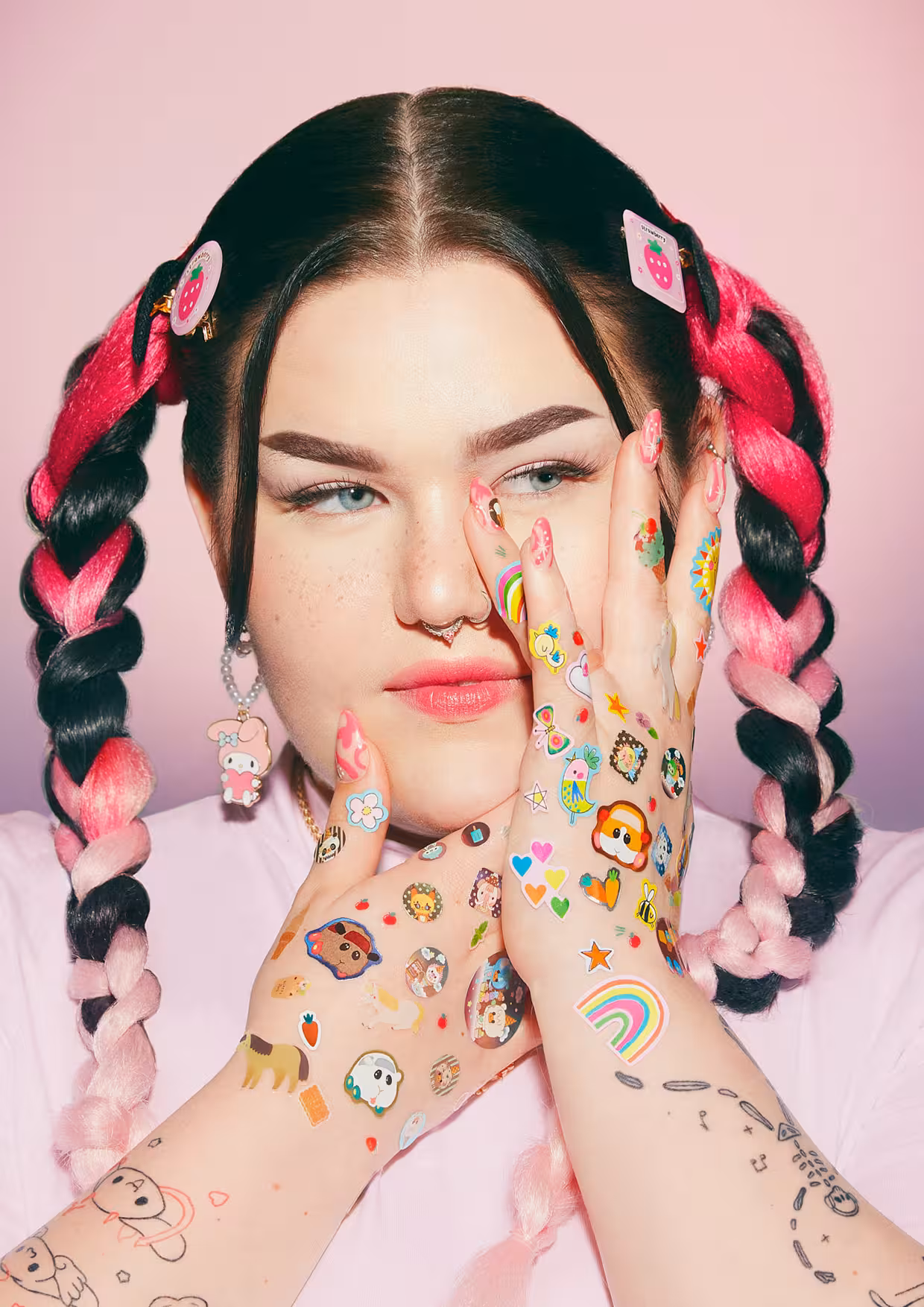 A close-up portrait of Mae Stephens with colourful braided hair, her hands decorated with small stickers framing her face.