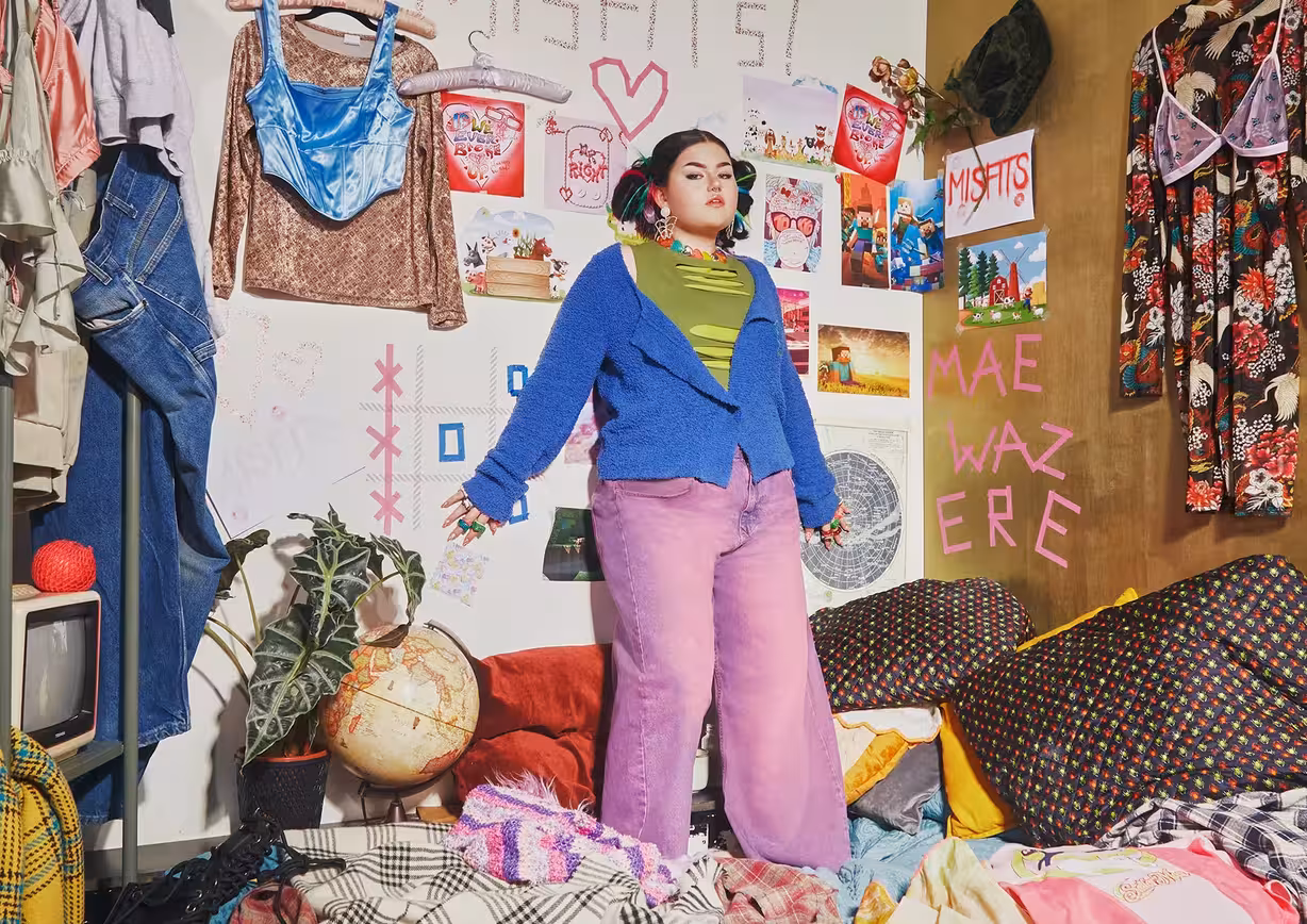 Artist Mae Stephens standing in a creatively cluttered room, wearing a colourful outfit with a blue jacket and pink trousers.