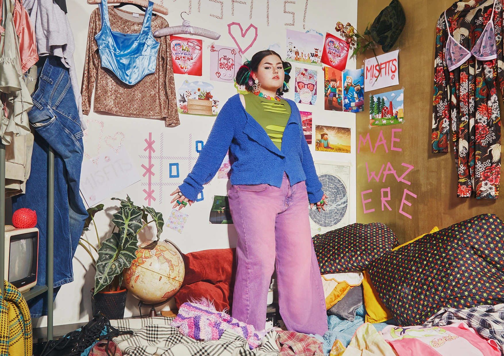 Artist Mae Stephens standing in a creatively cluttered room, wearing a colourful outfit with a blue jacket and pink trousers.