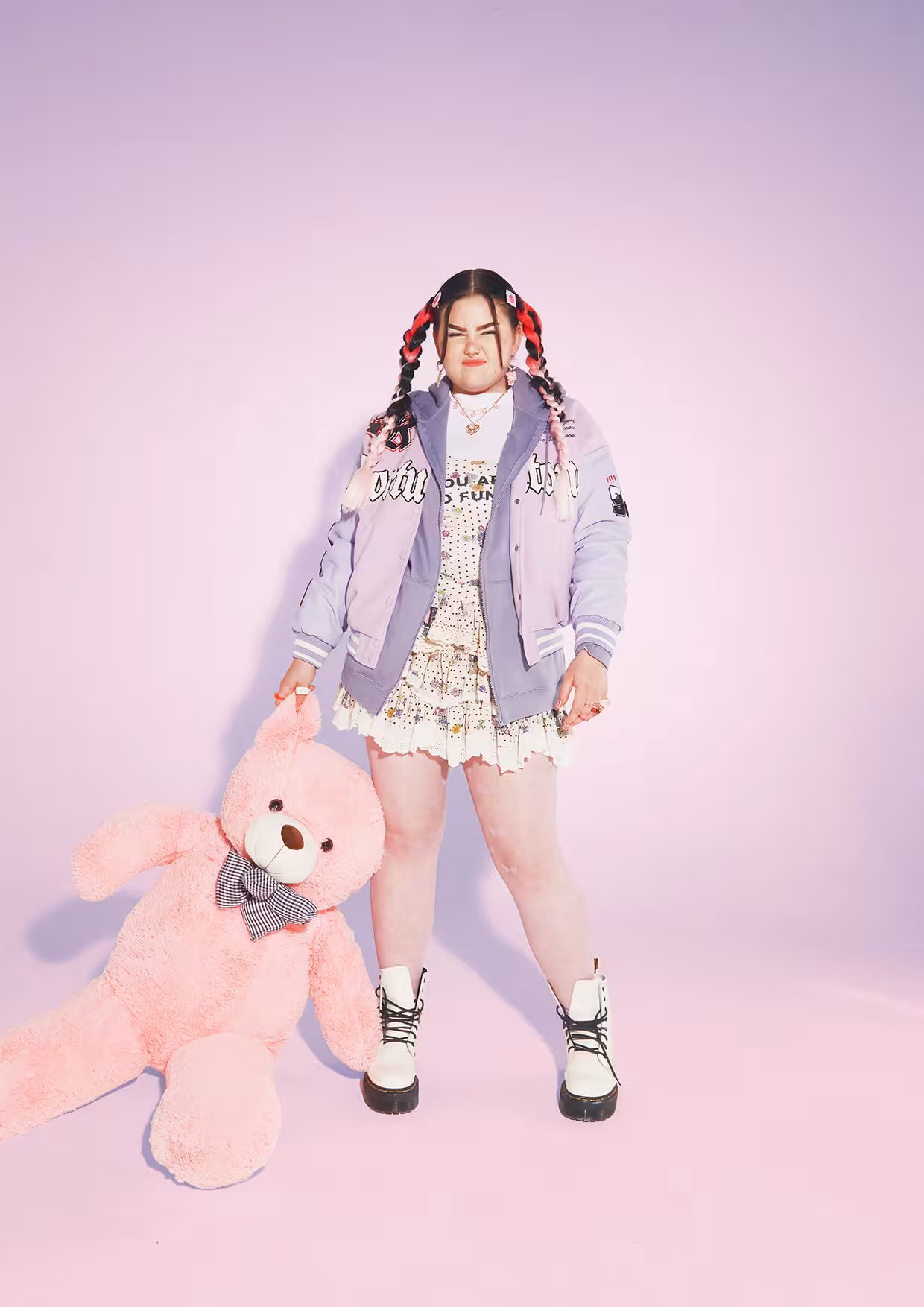 Mae Stephens standing against a pink background, wearing a pastel jacket and holding a pink teddy bear.
