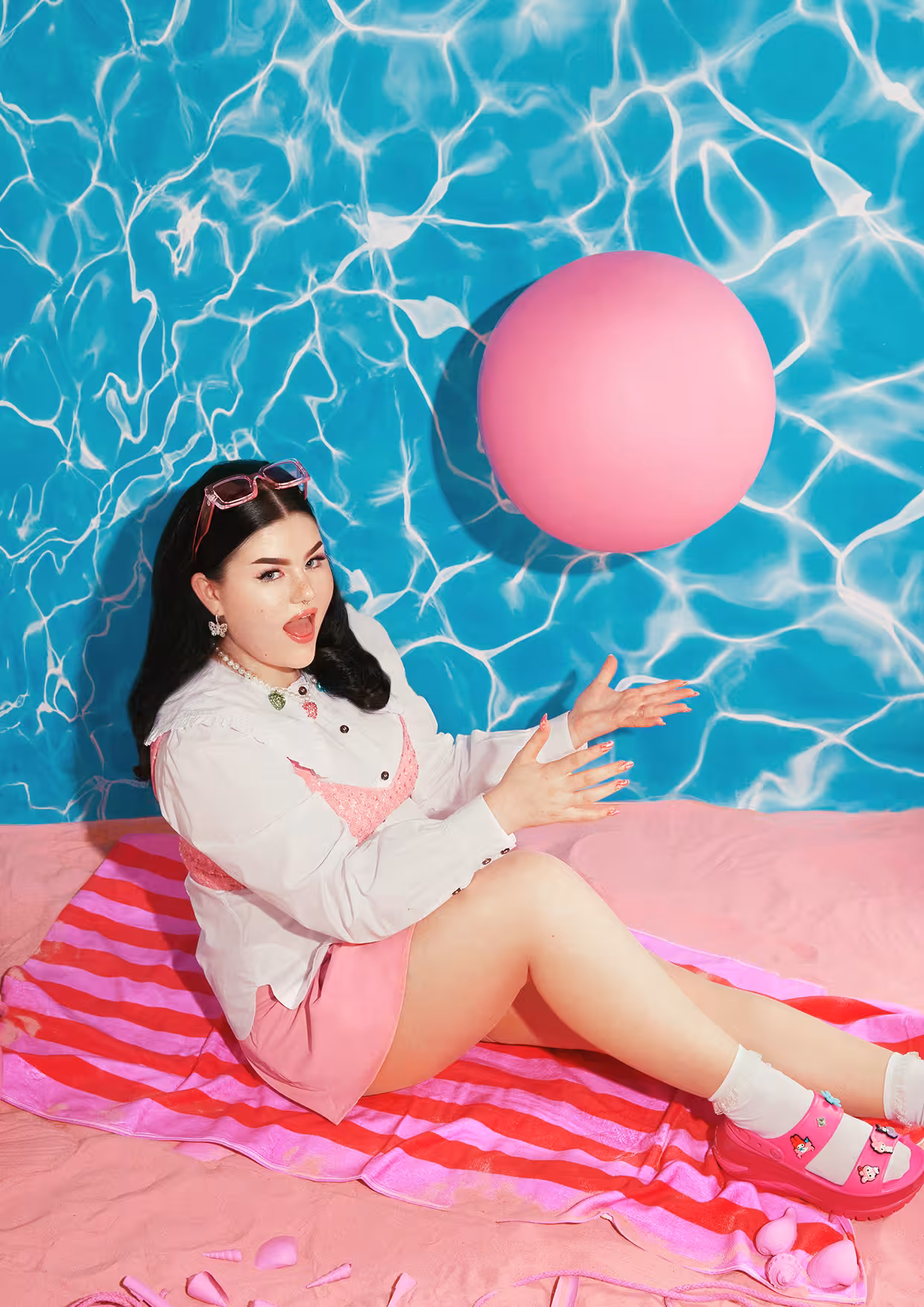 Mae Stephens lying on a pink towel on pink sand next to a bright blue swimming pool background, looking towards a pink ball.