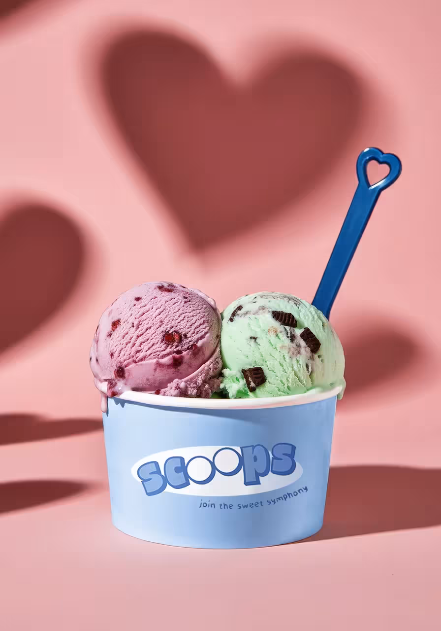 Pink and green scoops in a blue ice cream cup with a heart-shaped spoon against a pink background with a heart shadow.